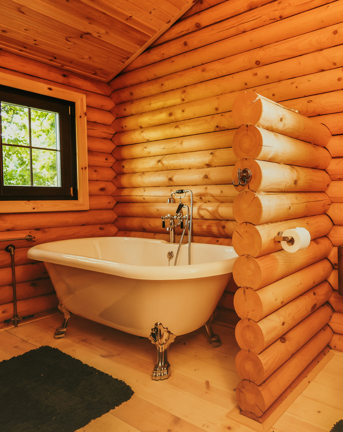 ErveHilbrand Rustieke badkamer in een houten blokhut met vrijstaand bad op pootjes, houten wanden en vloer, een raam met uitzicht op groen, en een houten scheidingsmuur met toiletrolhouder.