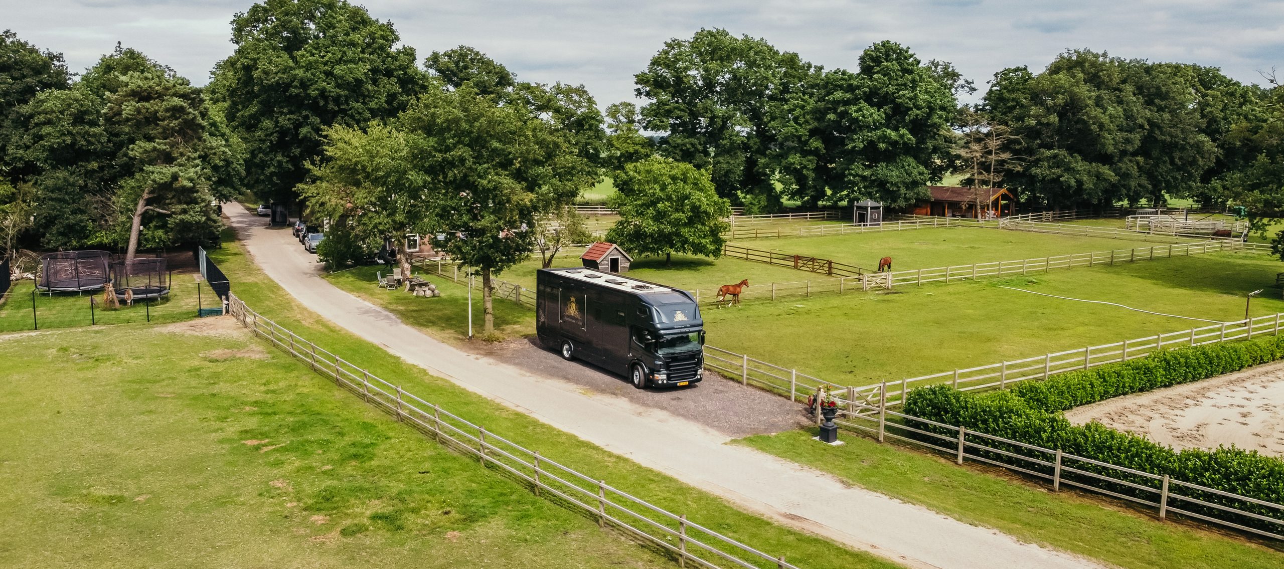 FotoOnderin Luchtfoto van een paardenaccommodatie met een grote zwarte paardenvrachtwagen geparkeerd langs een oprijlaan, omringd door weilanden met grazende paarden, een buitenrijbaan en houten omheiningen tussen groene bomen en gebouwen.