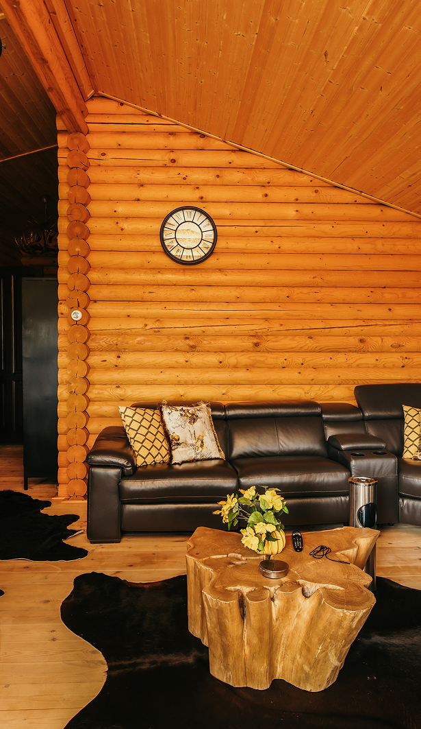 Recreatie-Bank Sfeervolle woonkamer in een blokhut met houten wanden en plafond, een zwarte leren hoekbank met sierkussens, een klok aan de muur en een robuuste boomstam-salontafel met bloemen en afstandsbediening.