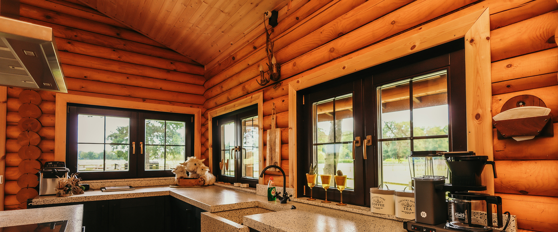 Sfeergroot Houten keukeninterieur van een blokhut met ramen die uitzicht bieden op een groene omgeving. Op het aanrecht staan een koffiezetapparaat, drie gouden glazen, en potten met opschriften ‘Coffee’ en ‘Tea’.