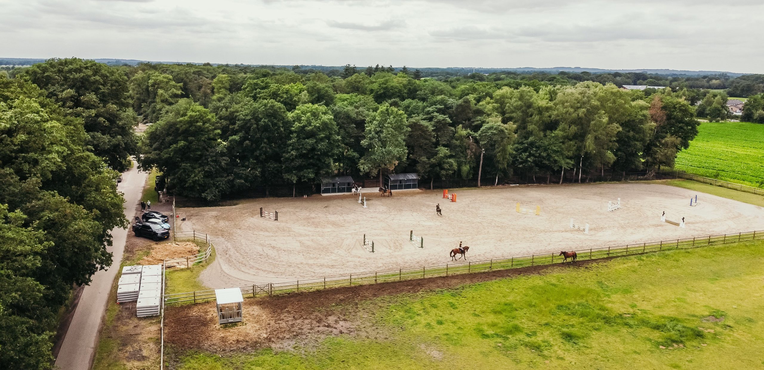 horse-power Luchtfoto van een grote buitenrijbaan omringd door bomen, waarop meerdere ruiters met hun paarden aan het trainen zijn tussen springhindernissen. Langs de kant staan enkele auto’s en toeschouwers, met weilanden en bos op de achtergrond.
