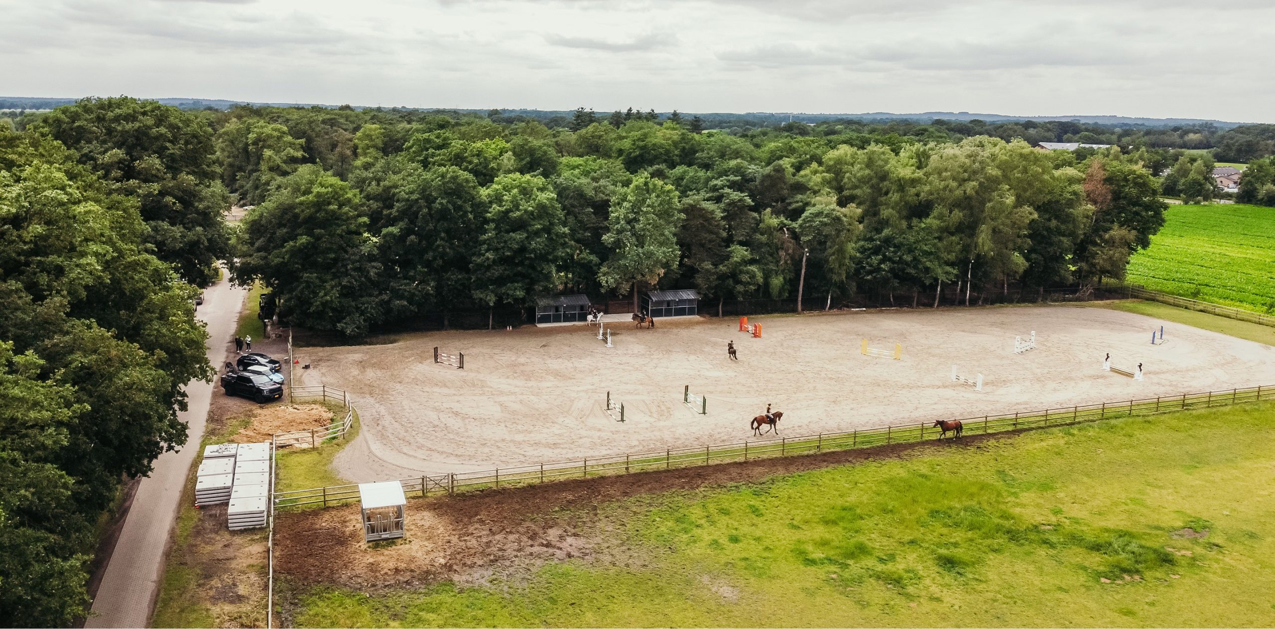 horse-power Luchtfoto van een grote buitenrijbaan omringd door bomen, waarop meerdere ruiters met hun paarden aan het trainen zijn tussen springhindernissen. Langs de kant staan enkele auto’s en toeschouwers, met weilanden en bos op de achtergrond.