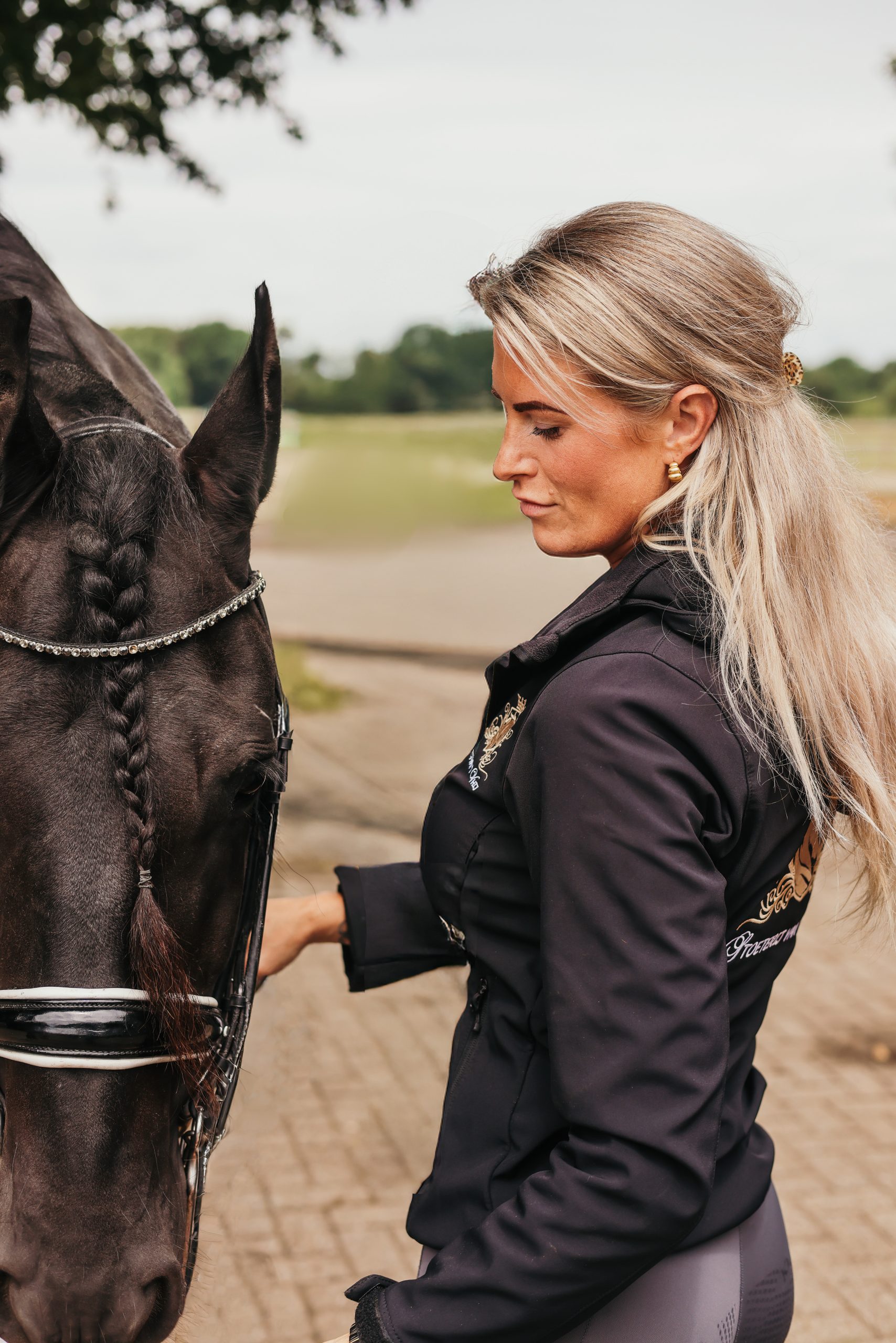 Vrouw met lang blond haar in paardrijkleding staat naast een zwart paard met gevlochten manen en hoofdstel, op een erf met weiland op de achtergrond.