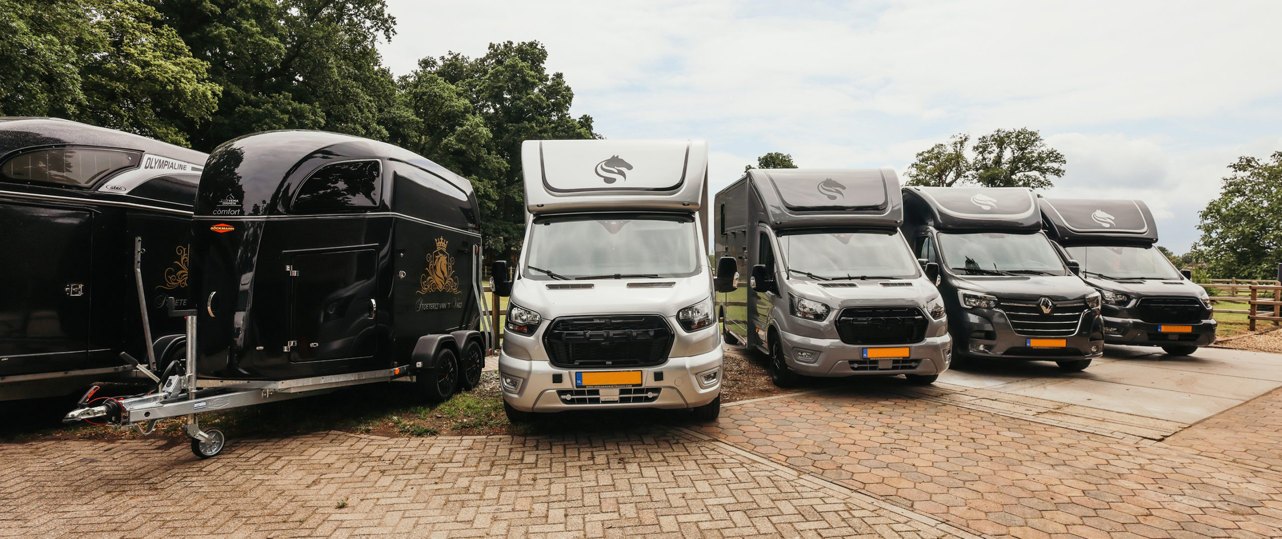 Rij van luxe zwarte en grijze paardenvrachtwagens en trailers, geparkeerd op een erf met bomen op de achtergrond.